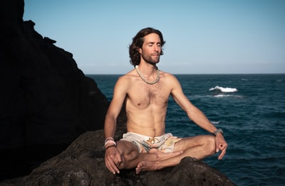白天，穿着棕色短裤的赤裸上身男子坐在海边的岩石上